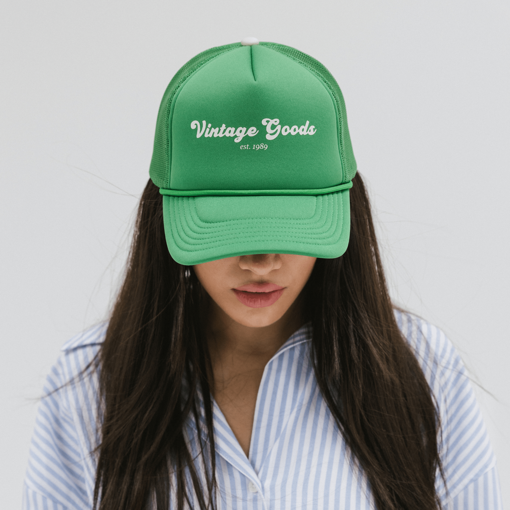 Woman wearing a vintage green colored trucker hat with 'Vintage Goods' text and a white shirt with blue lines, looking down against a plain background #color_vintage green