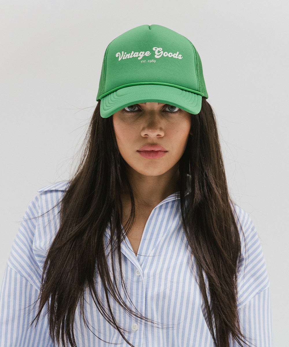 Woman wearing a vintage green colored trucker hat with 'Vintage Goods' text and a white shirt with blue lines against a plain background #color_vintage green
