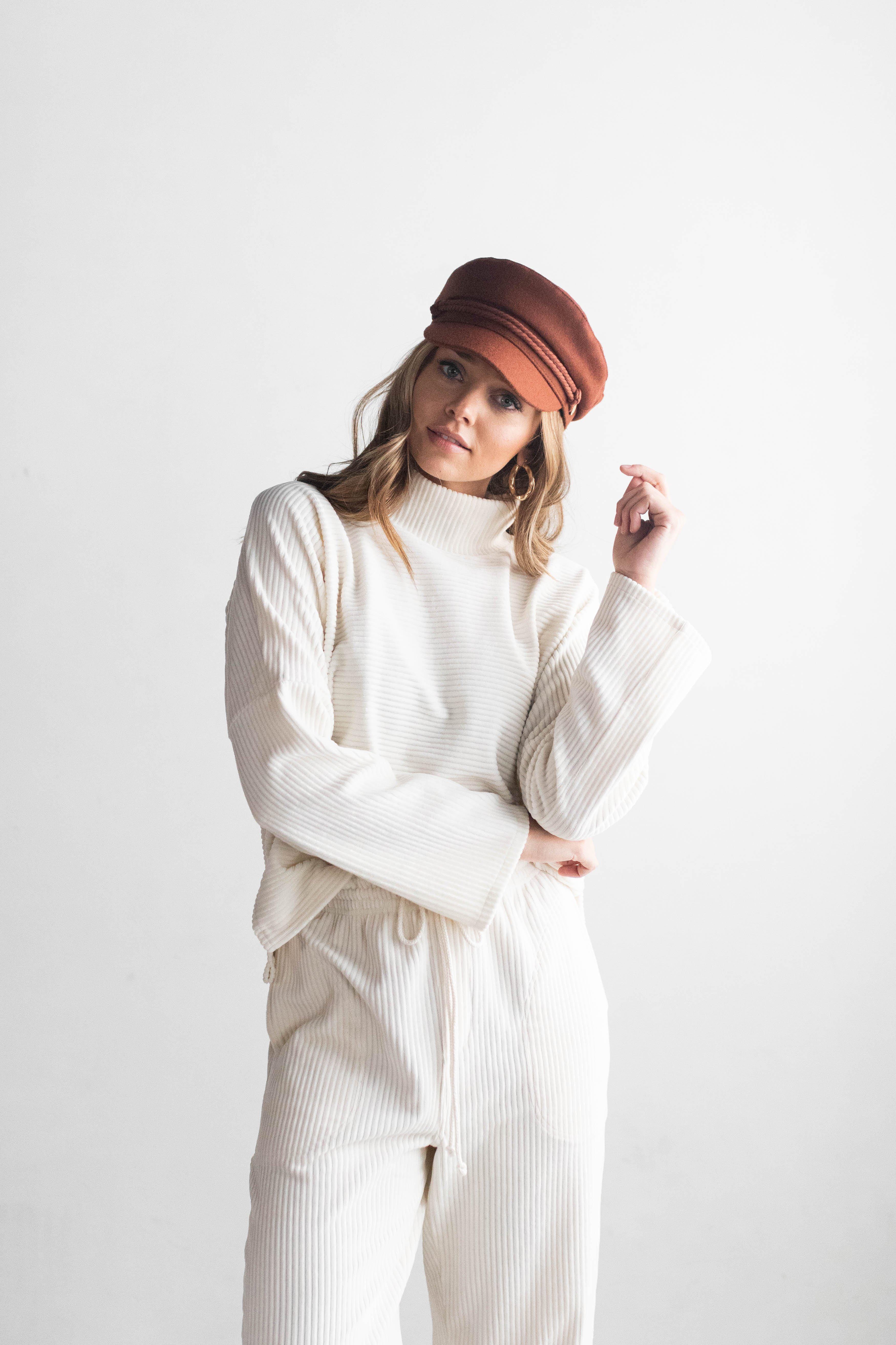 Woman wearing a chocolate brown colored fisherman cap with a rope detail, a white sweater and white pants against a plain background #color_chocolate brown
