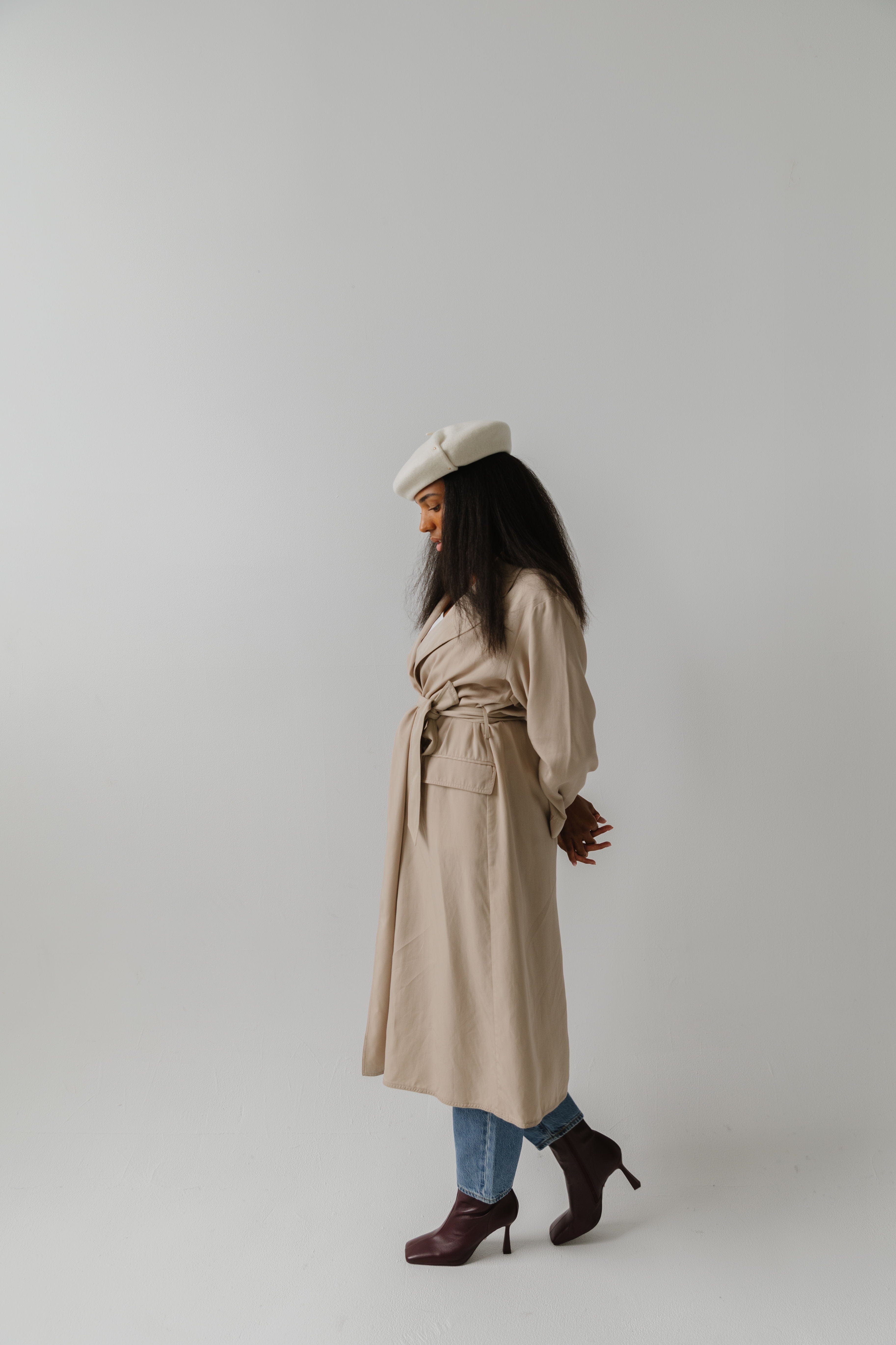 Woman wearing an off white beret, a beige coat, jeans and brown boots, posing against a plain background #color_off white