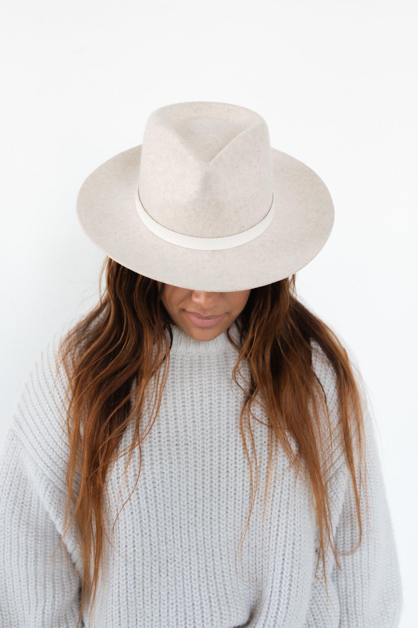 Woman wearing an ivory colored hat with an off white leather band with tassels and a white sweater, looking down against a plain background #color_off white