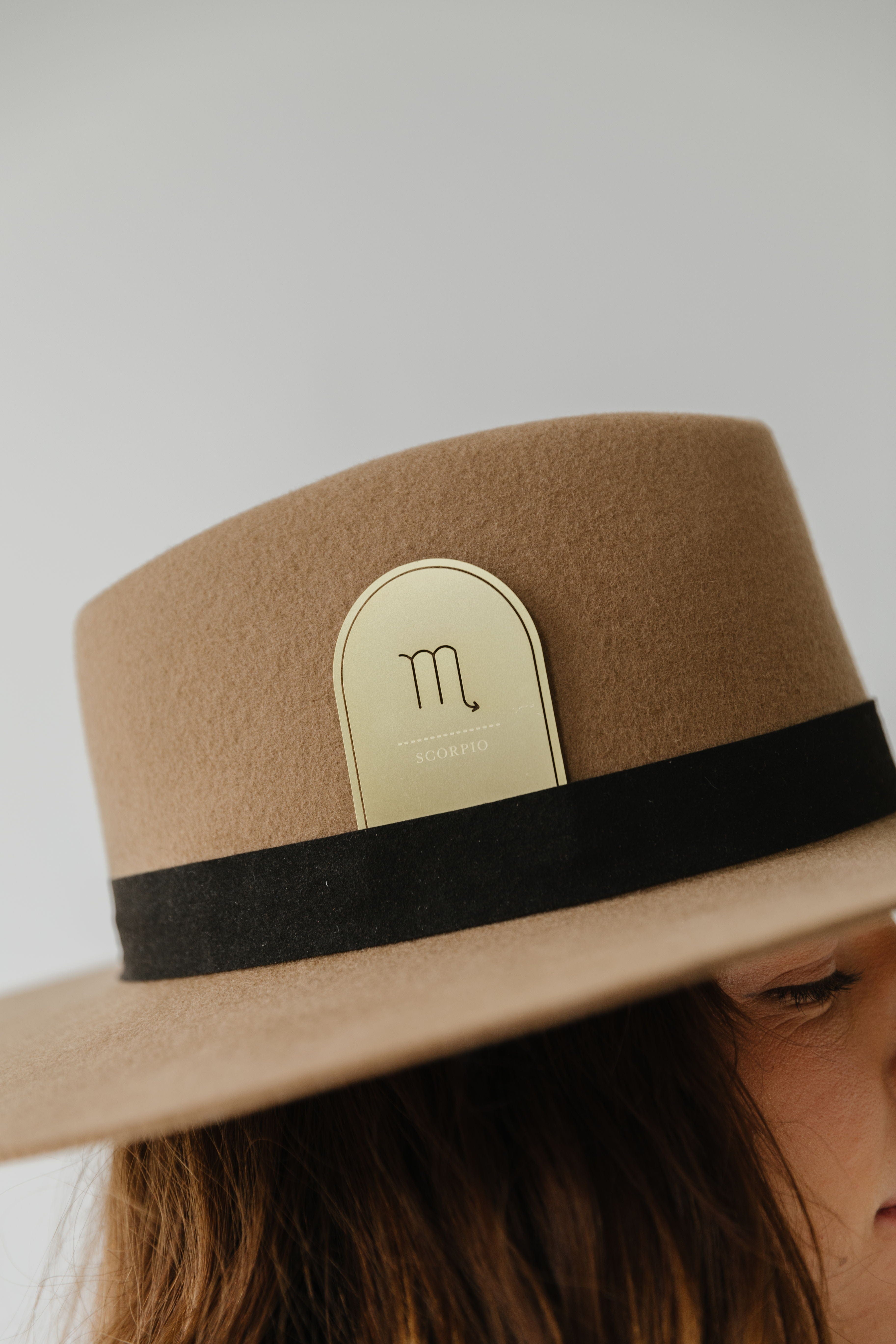 Woman wearing a brown hat with a black band and a gold 'Scorpio' zodiac band card against a plain background #color_gold