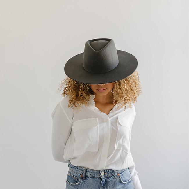 Woman wearing a dark grey fedora hat with a triangle crown, a white shirt and blue jeans against a plain background #color_dark grey
