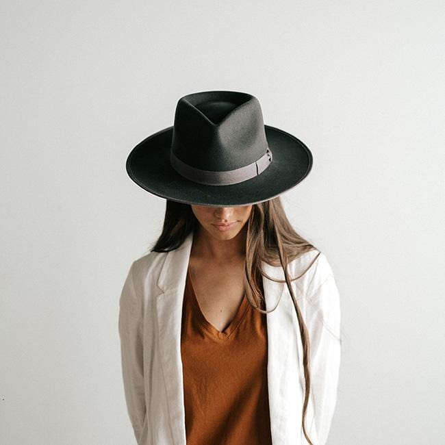 Woman wearing a dark grey fedora hat with a band and a brown t-shirt with a white jacket, looking down against a plain background #color_dark grey