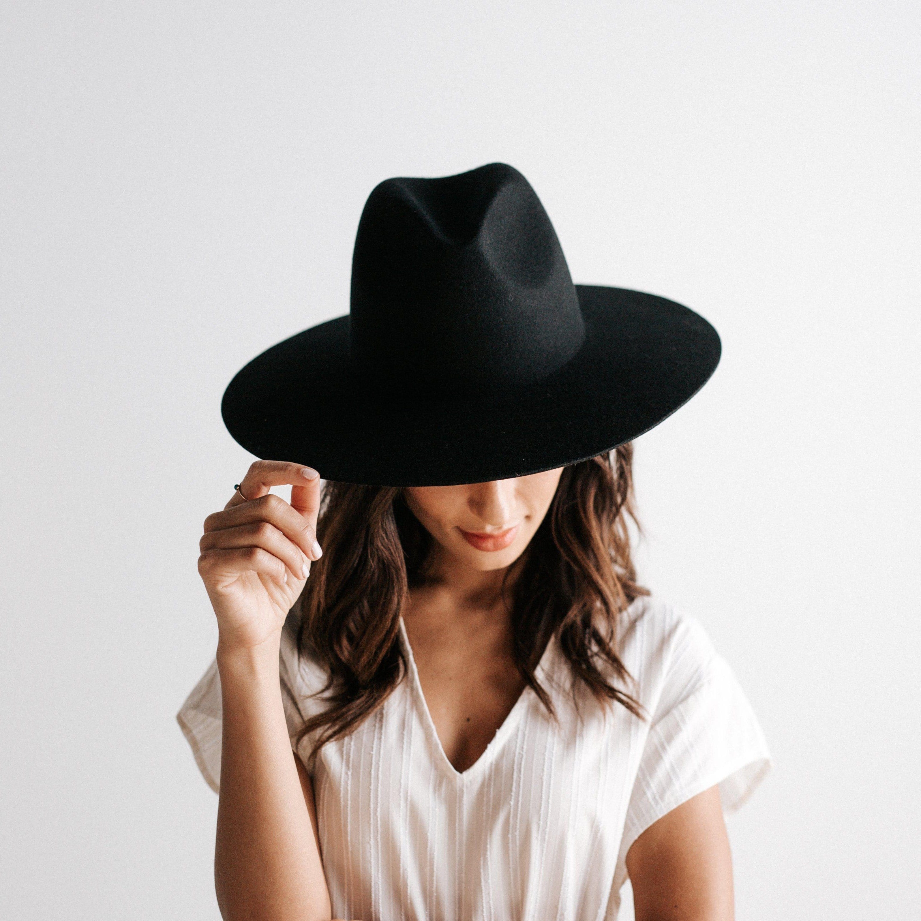 Woman wearing a black wide brim fedora hat and a white top against a plain background #color_black