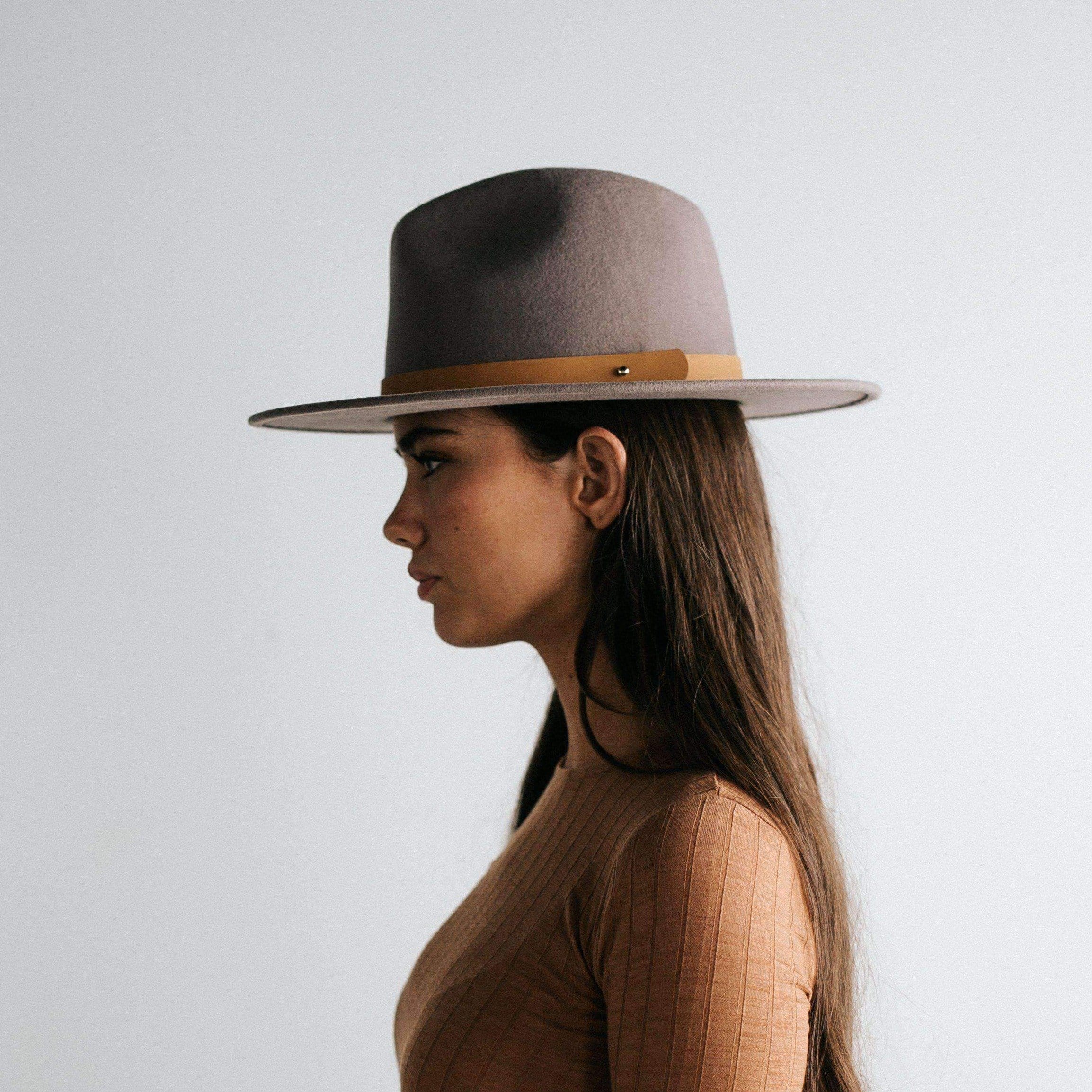 Woman wearing a light grey fedora hat with a hat band and a brown top, facing sideways against a plain background #color_light grey
