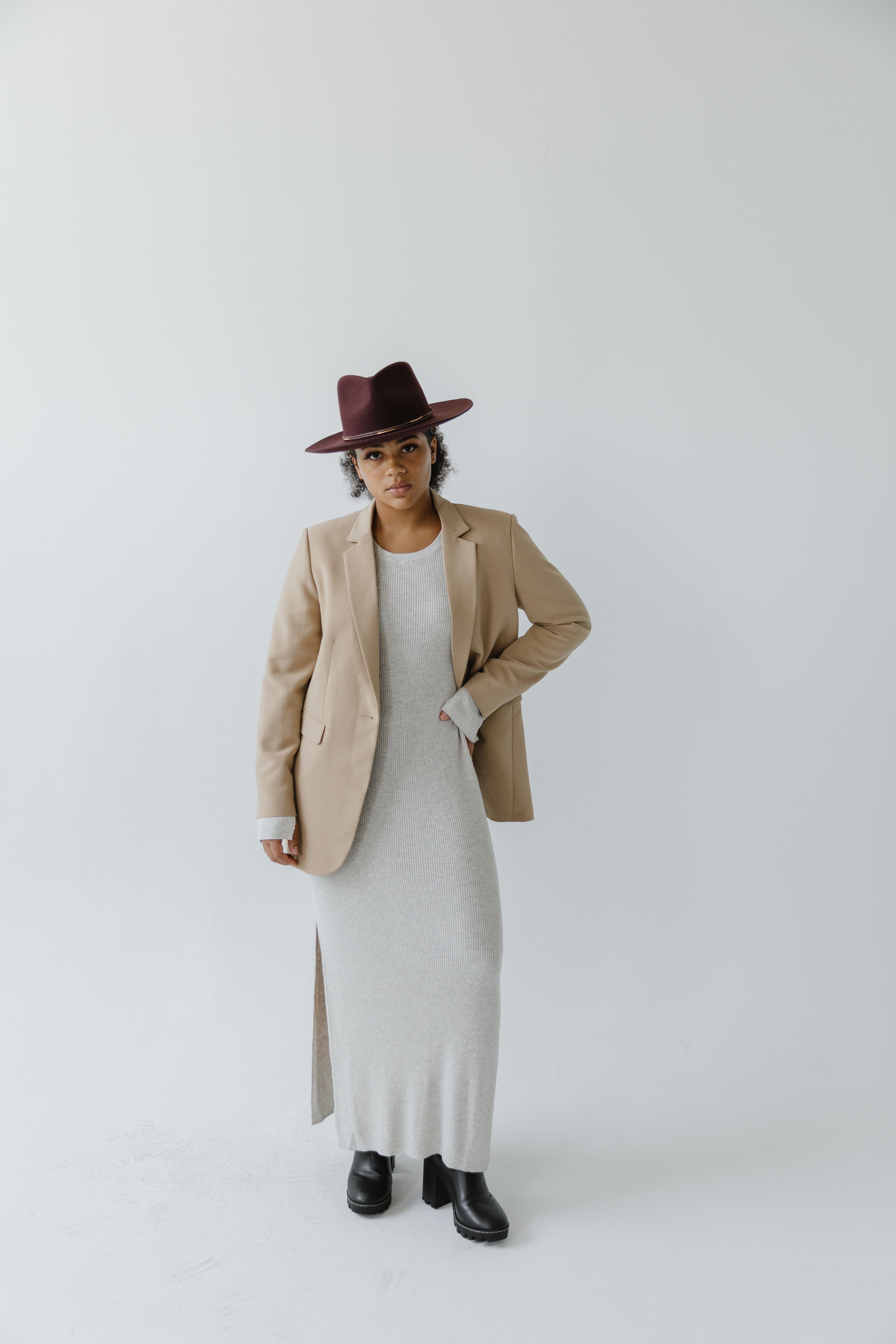 Woman wearing a dark cherry colored fedora hat, a white dress with a beige blazer and black boots against a plain background #color_dark cherry