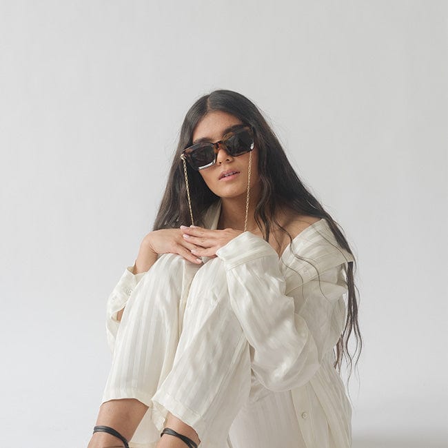 Woman wearing sunglasses with a gold chain strap, a white shirt and white pants, sitting against a plain background #color_gold