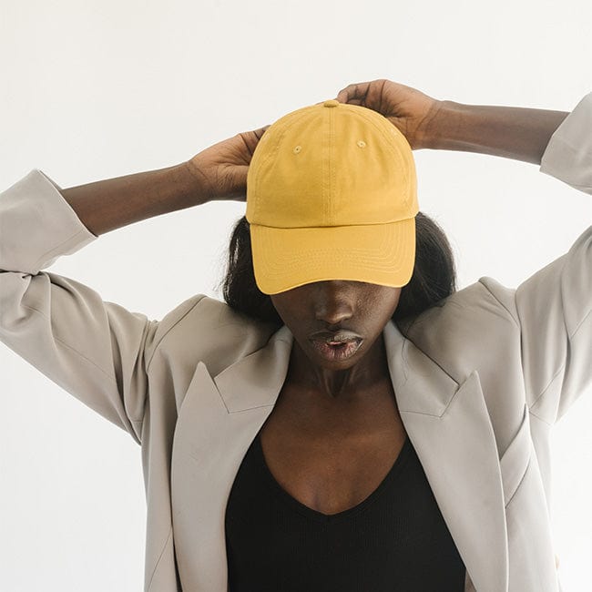 Woman wearing a mustard colored ball cap and a black top with a grey jacket against a plain background #color_mustard