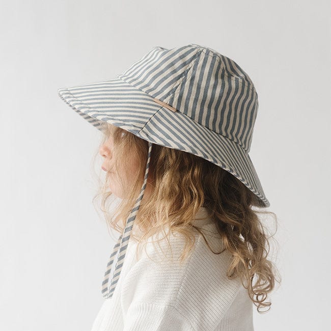 Young girl wearing a blue and white striped bucket hat and a white shirt, facing sideways against a plain background #color_striped-blue