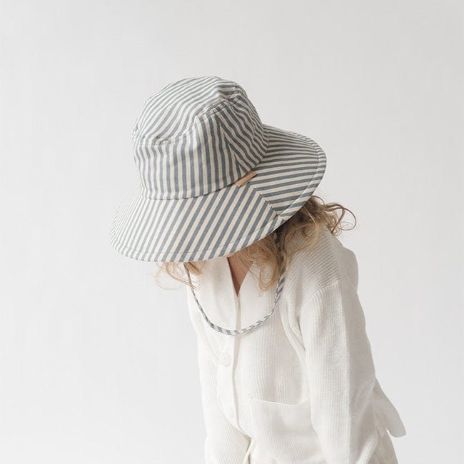 Young girl wearing a blue and white striped bucket hat and a white shirt, looking down against a plain background #color_striped-blue