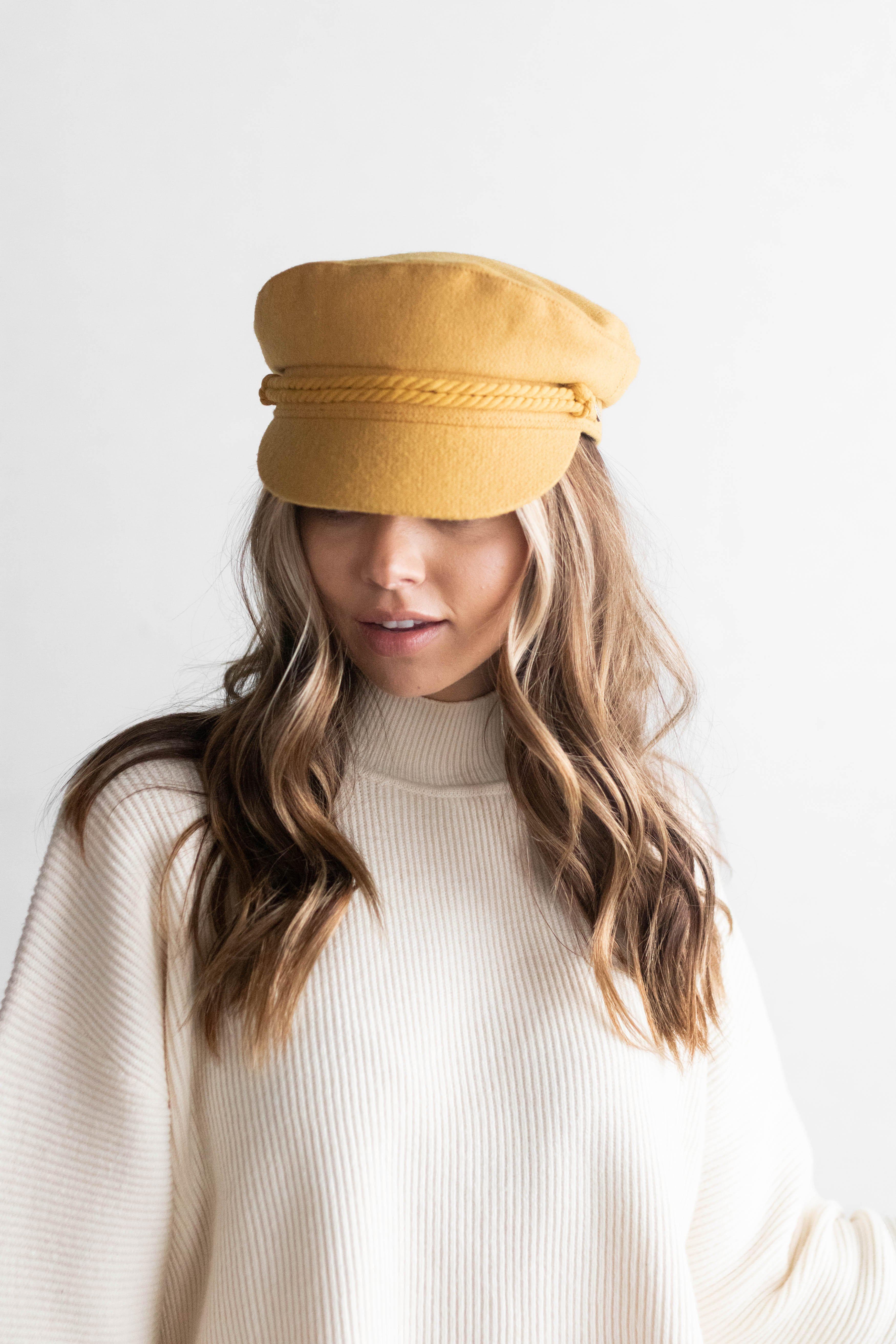 Woman wearing a mustard colored fisherman cap with a rope detail and a white sweater against a plain background #color_mustard