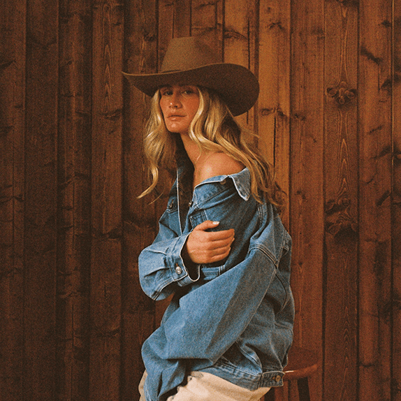 Woman wearing a cowboy hat and denim jacket against a wooden background