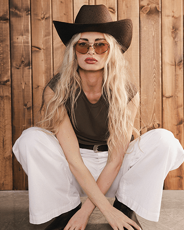Person wearing a cowboy hat and sunglasses sitting against a wooden wall.