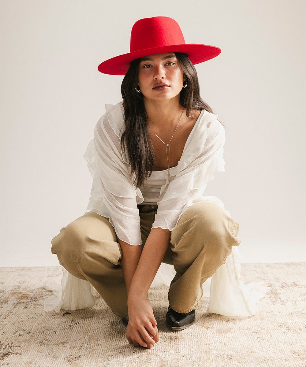 Woman wearing a cherry red felt hat, a white top with a white shirt, beige pants, and black shoes, sitting against a plain background #color_cherry red