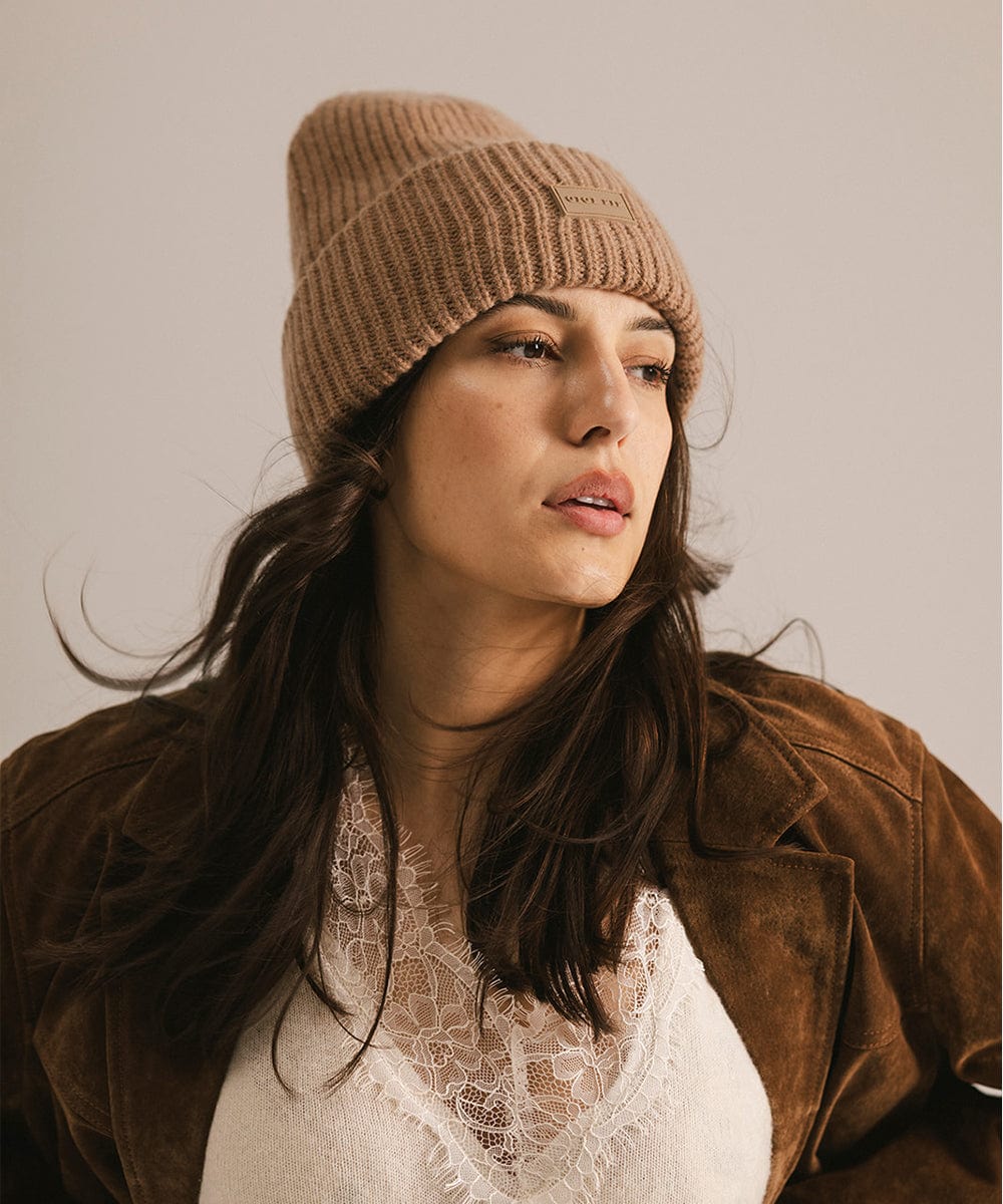 Woman wearing a brown knit beanie and brown jacket, looking sideways against a neutral background #color_brown