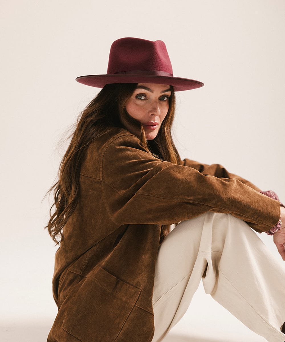 Woman wearing a merlot colored fedora hat with a band, a brown coat and white pants, sitting against a plain background #color_merlot