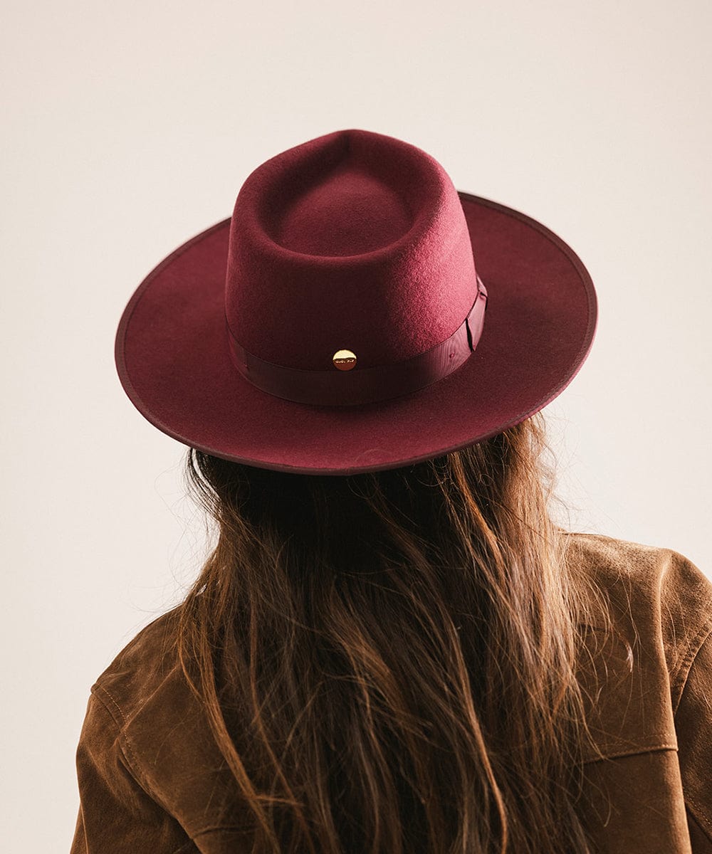Woman wearing a merlot colored fedora hat with a band and a brown coat, facing behind against a plain background #color_merlot