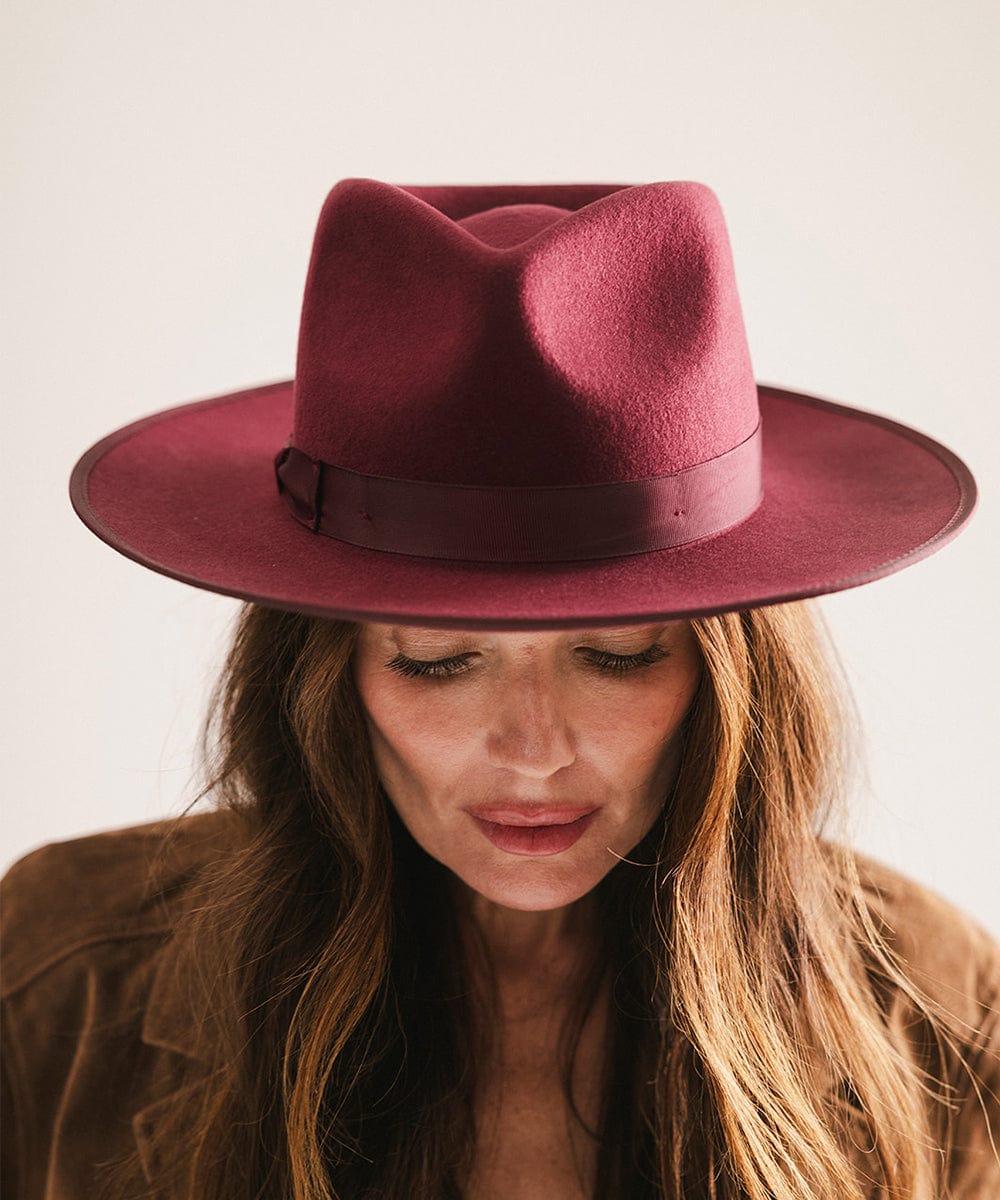 Woman wearing a merlot colored fedora hat with a band and a brown coat, looking down against a plain background #color_merlot