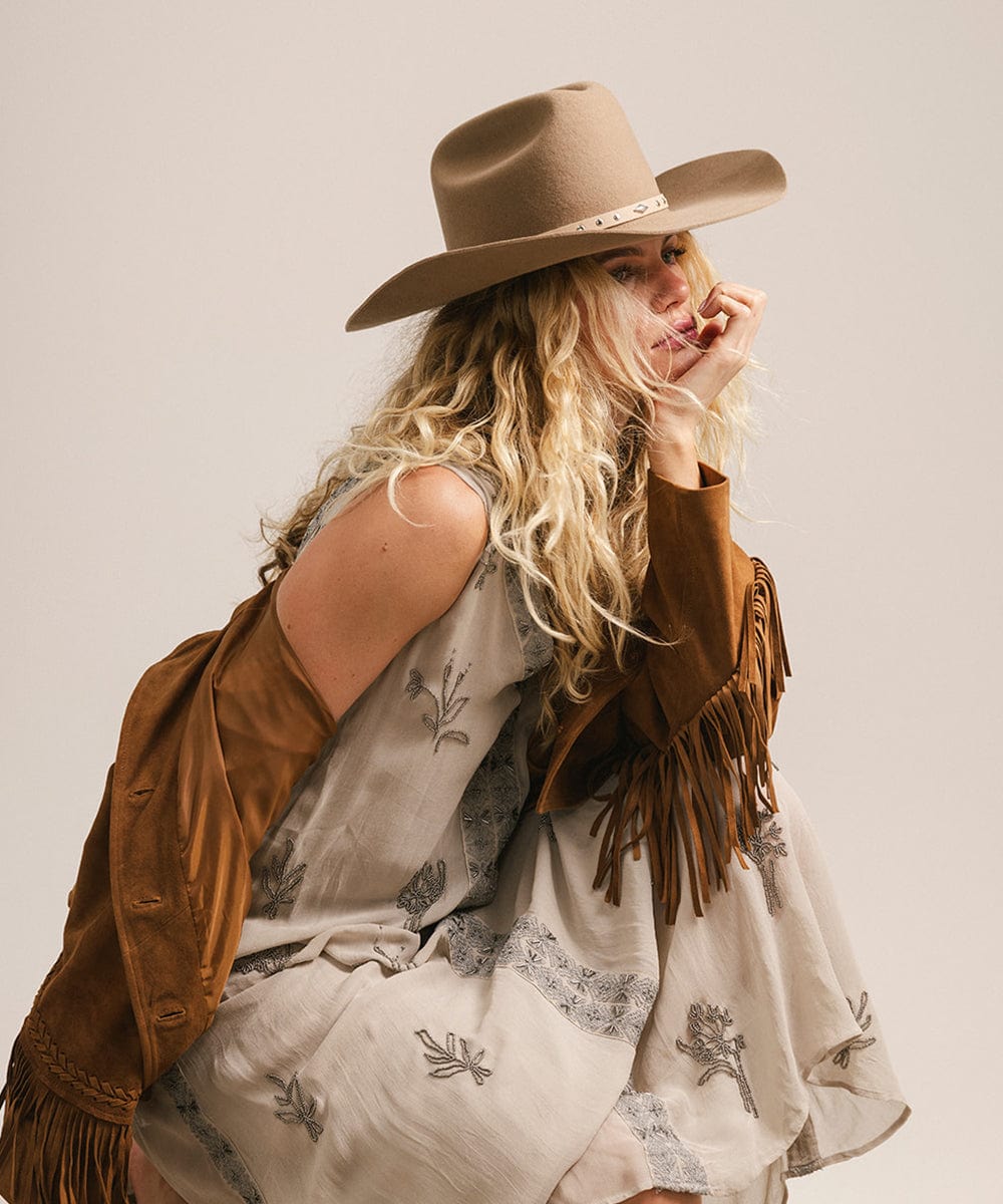 Woman wearing a brown cowboy hat with a hat band and a beige embroidered dress with a brown jacket, looking sideways against a plain background #color_brown