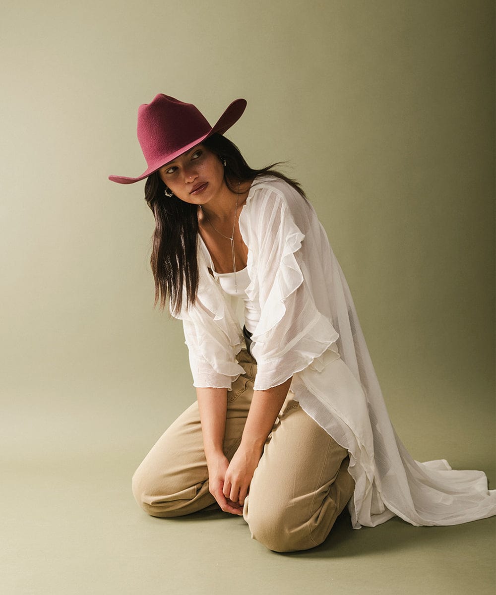 Woman wearing a merlot colored cowboy hat, a white blouse and beige pants, sitting against a plain background #color_merlot