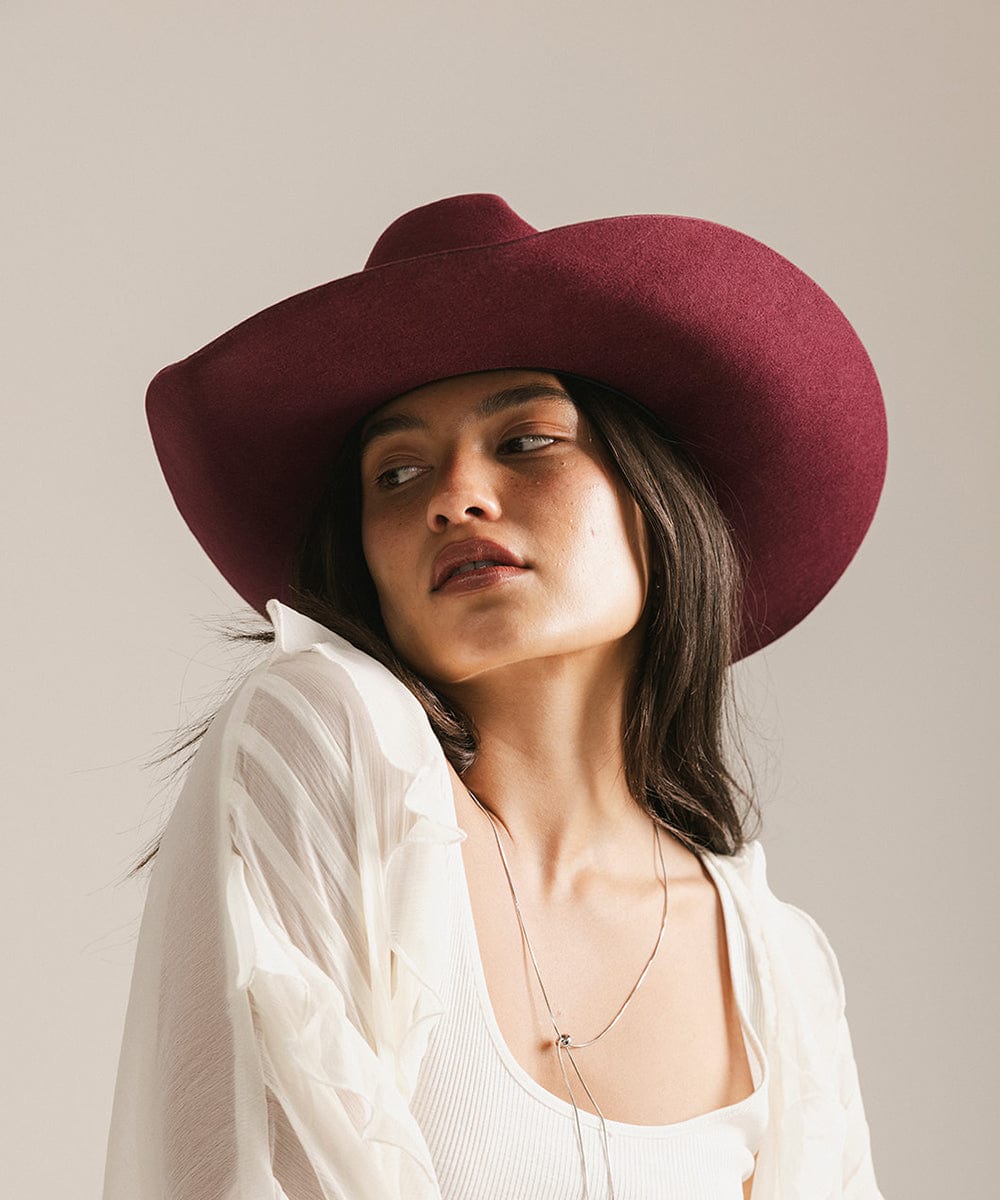 Woman wearing a merlot colored cowboy hat and a white top with a white shirt, looking sideways against a plain background #color_merlot