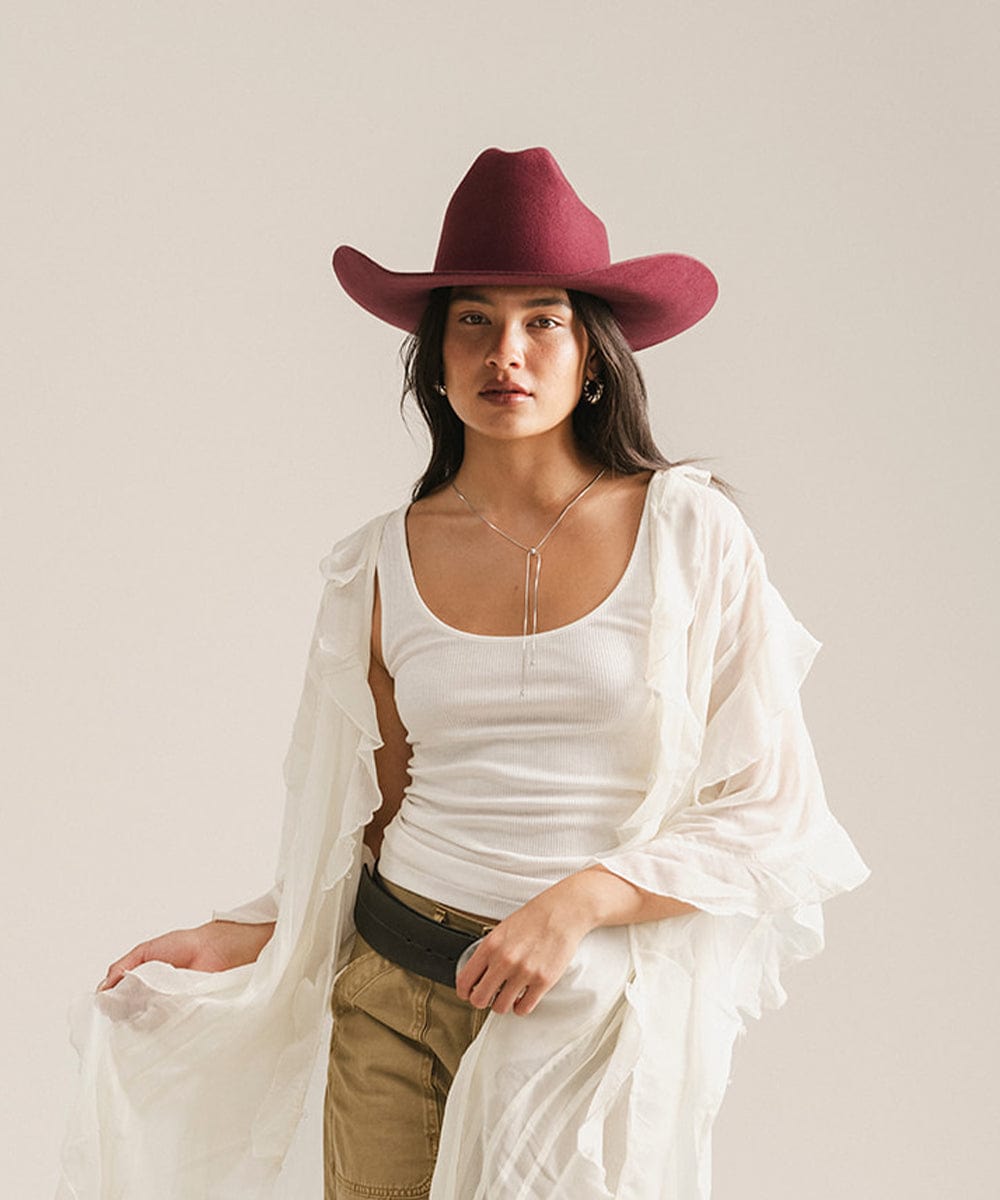 Woman wearing a merlot colored cowboy hat, a white top with a white shirt and beige pants against a plain background #color_merlot