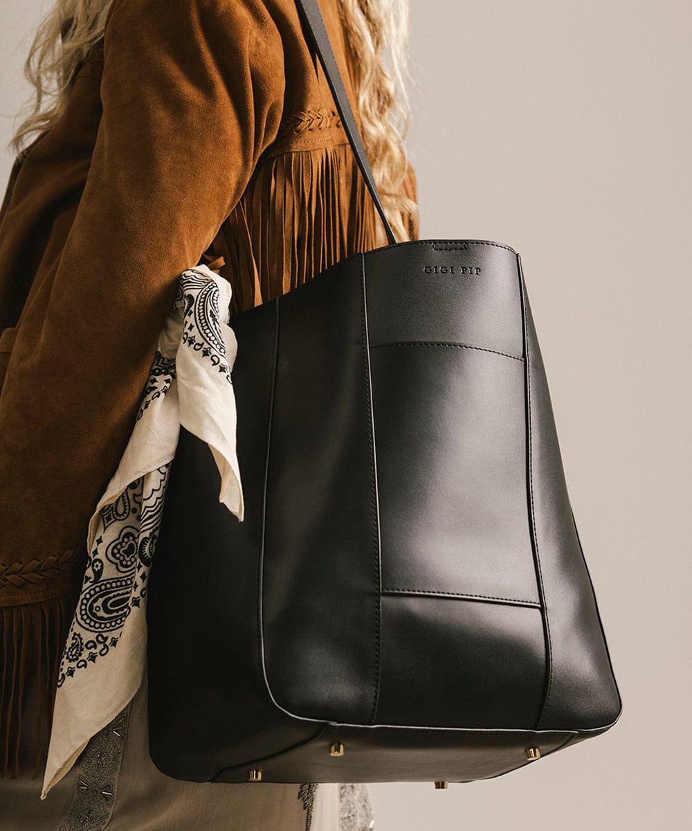 Black leather handbag held by a person wearing a brown jacket with fringe details. #color_black