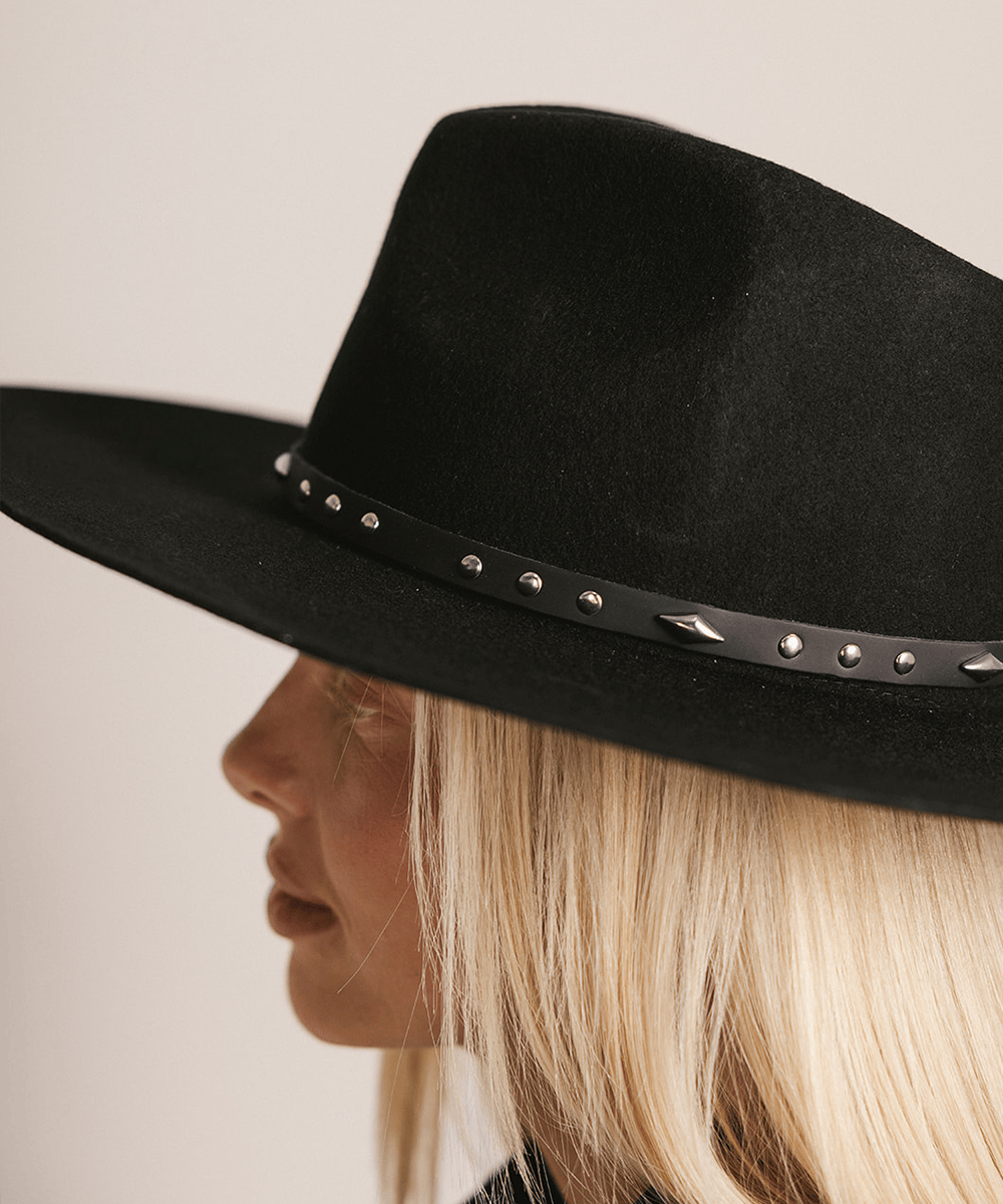 Woman wearing a black hat with a black studded leather hat band against a plain background #color_black