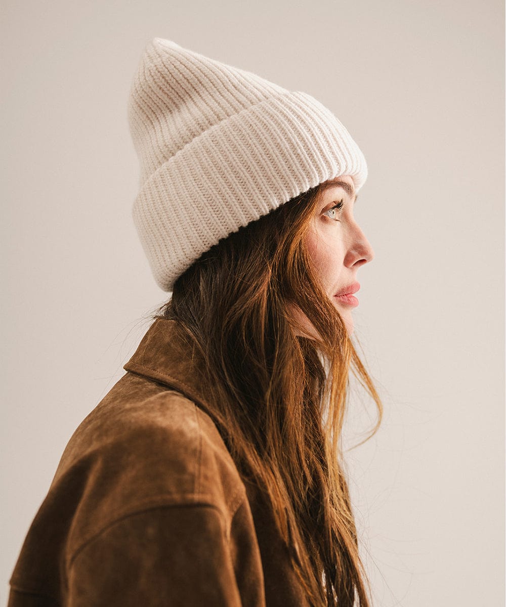 Woman wearing an off white knit beanie and brown jacket, facing sideways against a plain background #color_off white