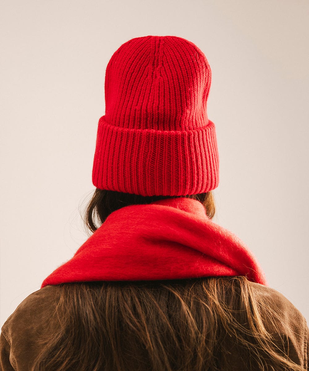 A woman wearing a ruby red colored ribbed beanie and scarf and a jacket, facing behind against a plain background #color_ruby red