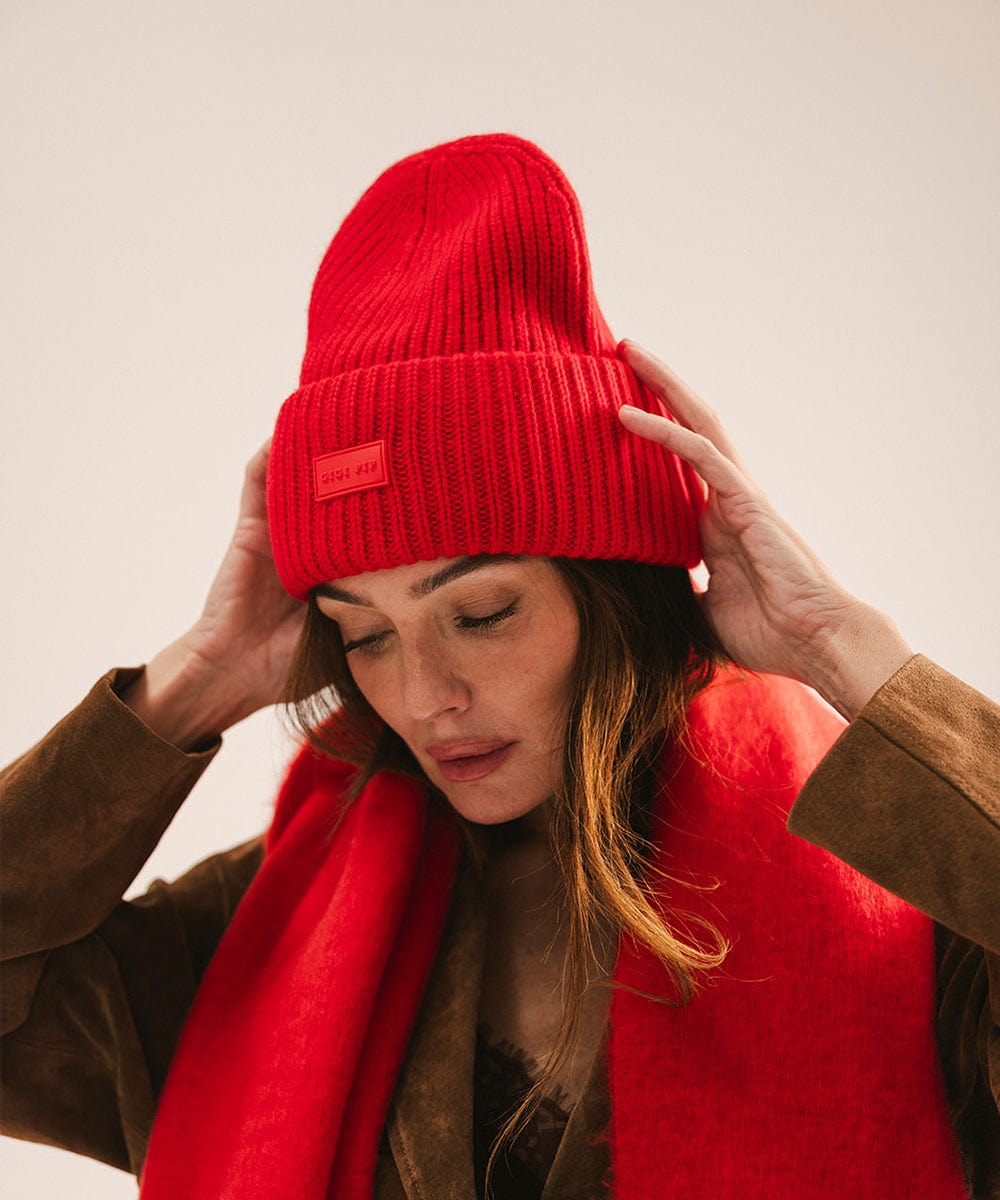 A woman wearing a ruby red colored ribbed beanie and scarf and a jacket, looking down against a plain background #color_ruby red