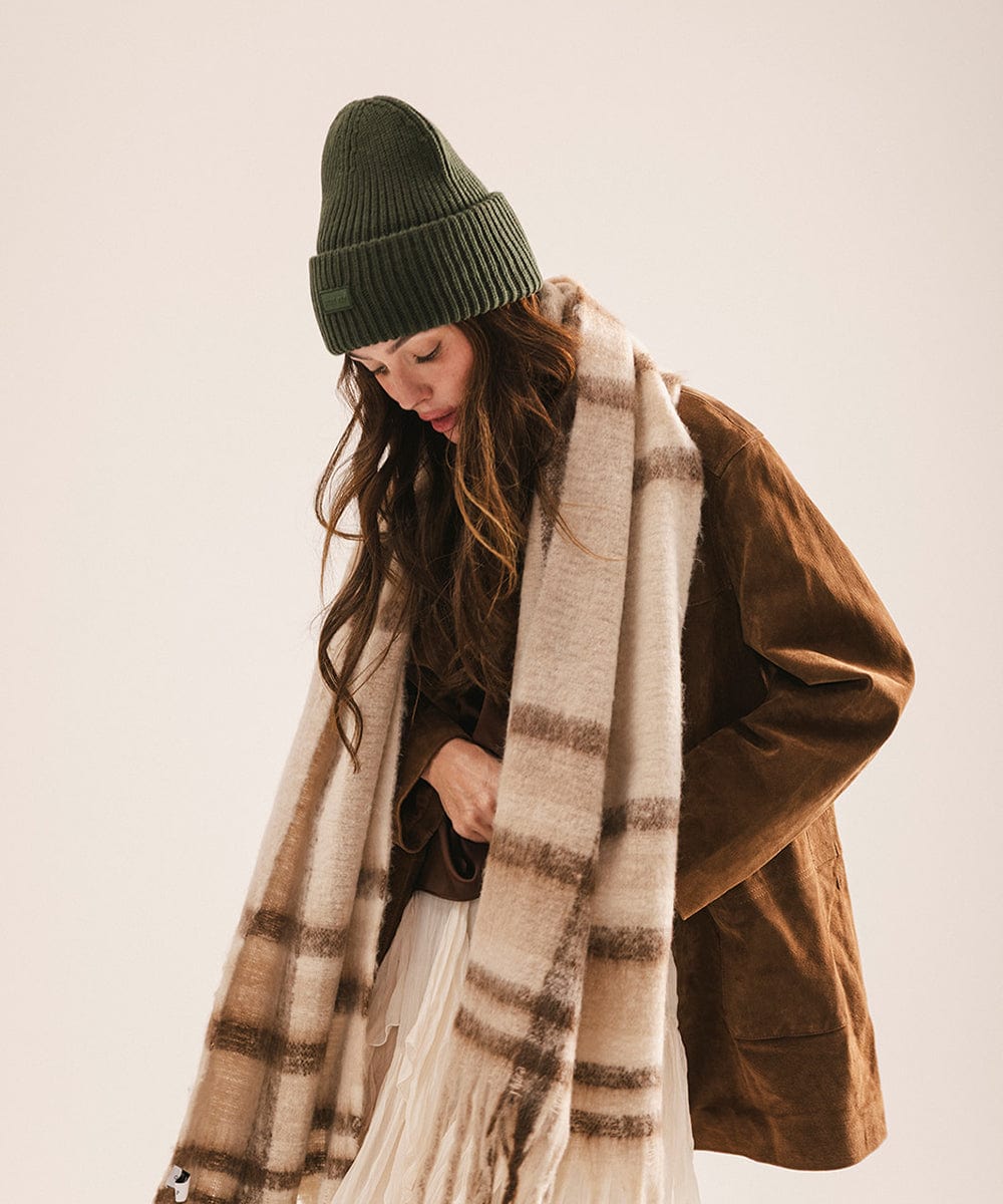 A woman wearing a dark green chunky ribbed beanie and a brown coat, looking down against a plain background #color_dark green