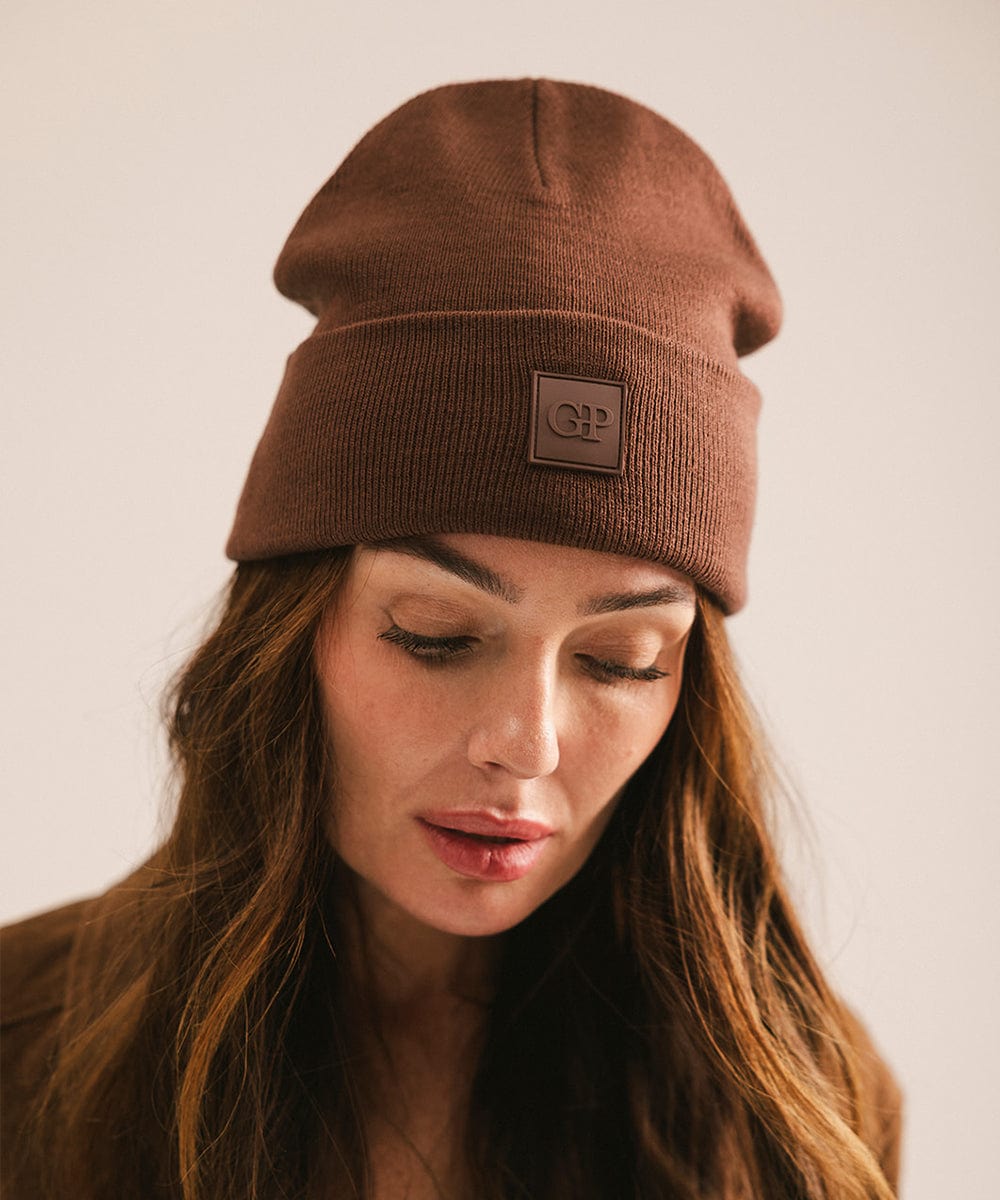 Woman wearing a chocolate colored beanie with a logo on a plain background #color_chocolate