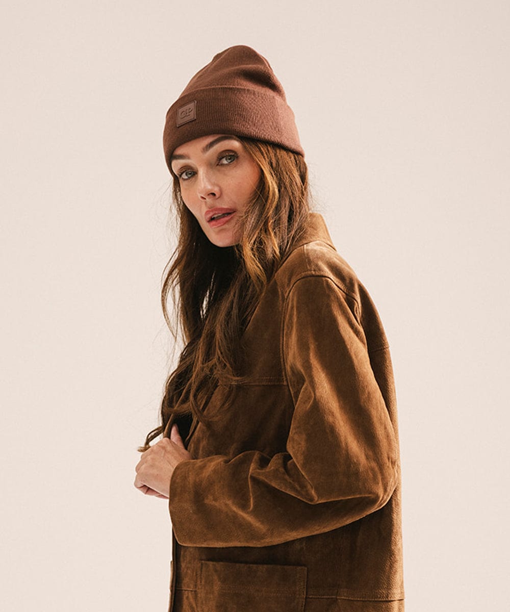 Woman wearing a chocolate colored beanie and brown jacket, posing sideways against a plain background #color_chocolate