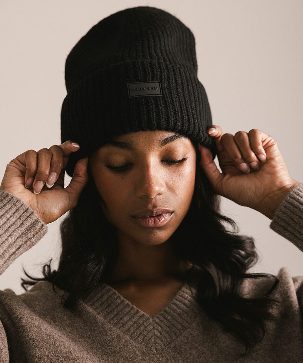 Woman wearing a black knit beanie with a brand logo against a neutral background #color_black