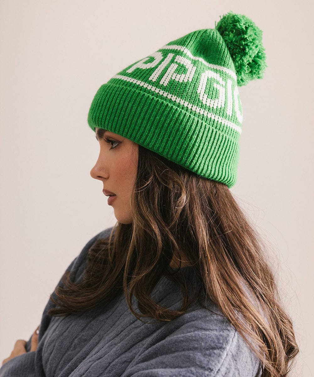 Woman wearing an evergreen colored knit beanie with pom-pom and 'GIGI PIP' branding and a blue sweater, facing sideways against a plain background #color_evergreen