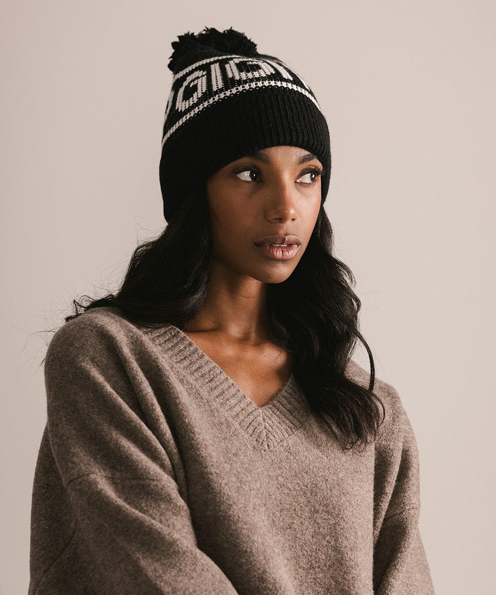 Woman wearing a black beanie with pom-pom and a beige sweater against a neutral background #color_black