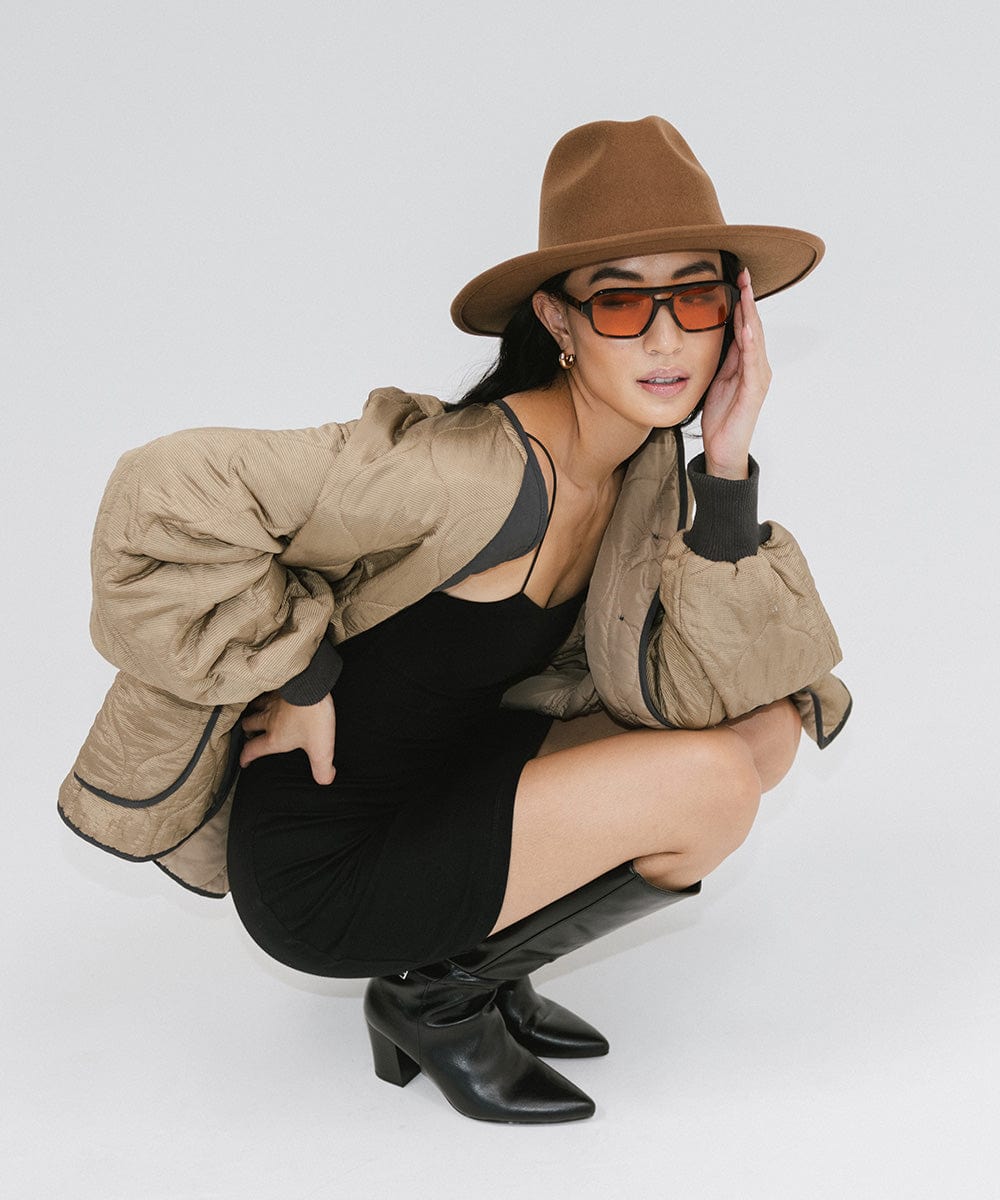 Woman wearing a brown fedora hat, sunglasses, a black top with a beige jacket and black boots against a plain background #color_brown