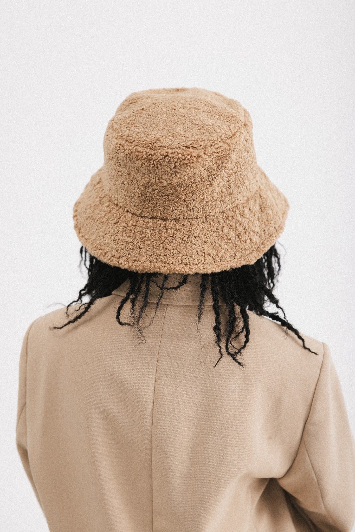 Person wearing a camel colored sherpa bucket hat and a beige coat  against a plain background #color_camel