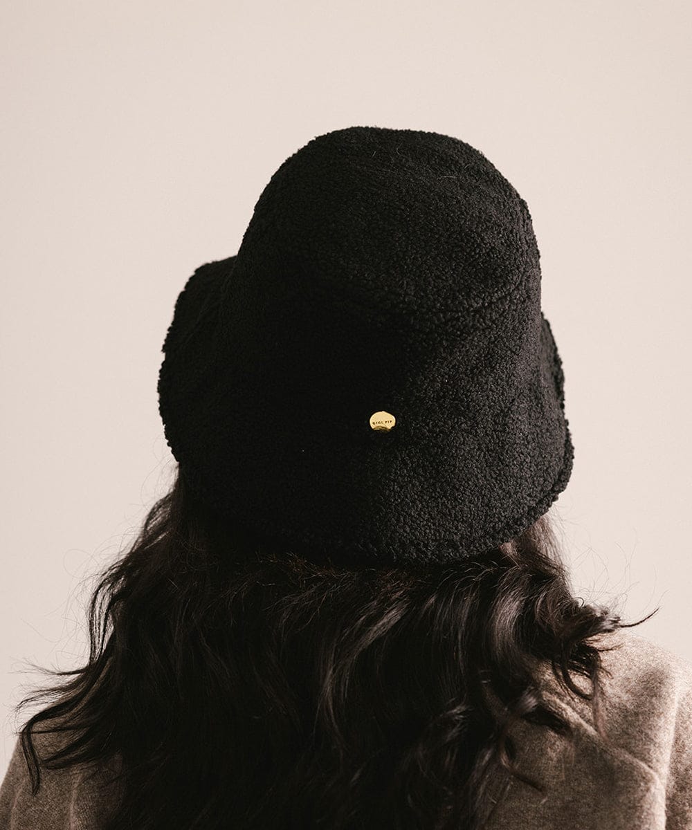 Person wearing a black sherpa bucket hat with a gold emblem on a plain background #color_black