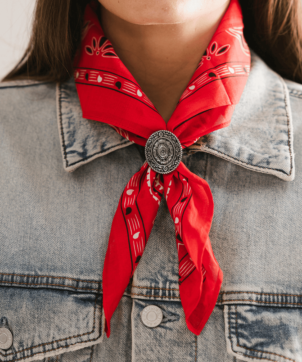 Person wearing a red patterned scarf tied around their neck with a Floral Oval Concho Bandana Slide and wearing a denim jacket. #color_silver