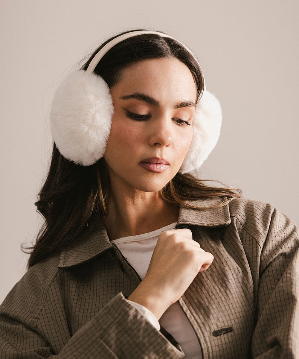 Woman wearing white fur ear muffs against a neutral background #color_winter white