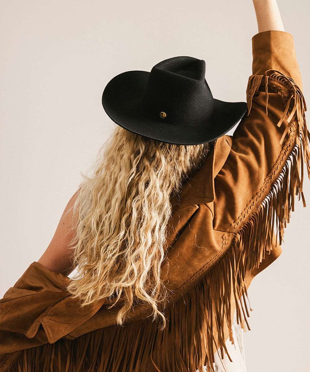 Person wearing a black cowboy hat and brown suede jacket with fringes on a neutral background #color_black