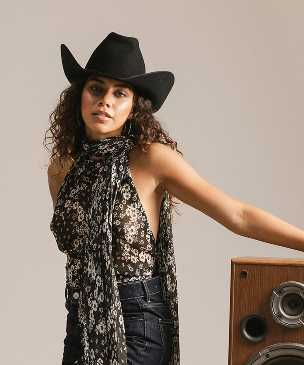 Woman wearing a black cowboy hat and floral scarf standing next to a wooden speaker. #color_black