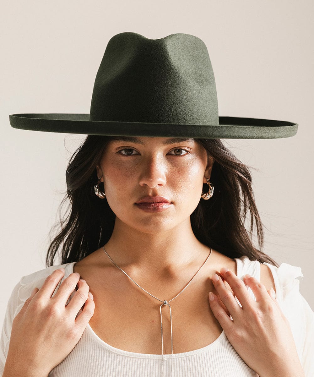 Woman wearing a dark green pencil brim fedora hat and a white top against a plain background #color_dark green
