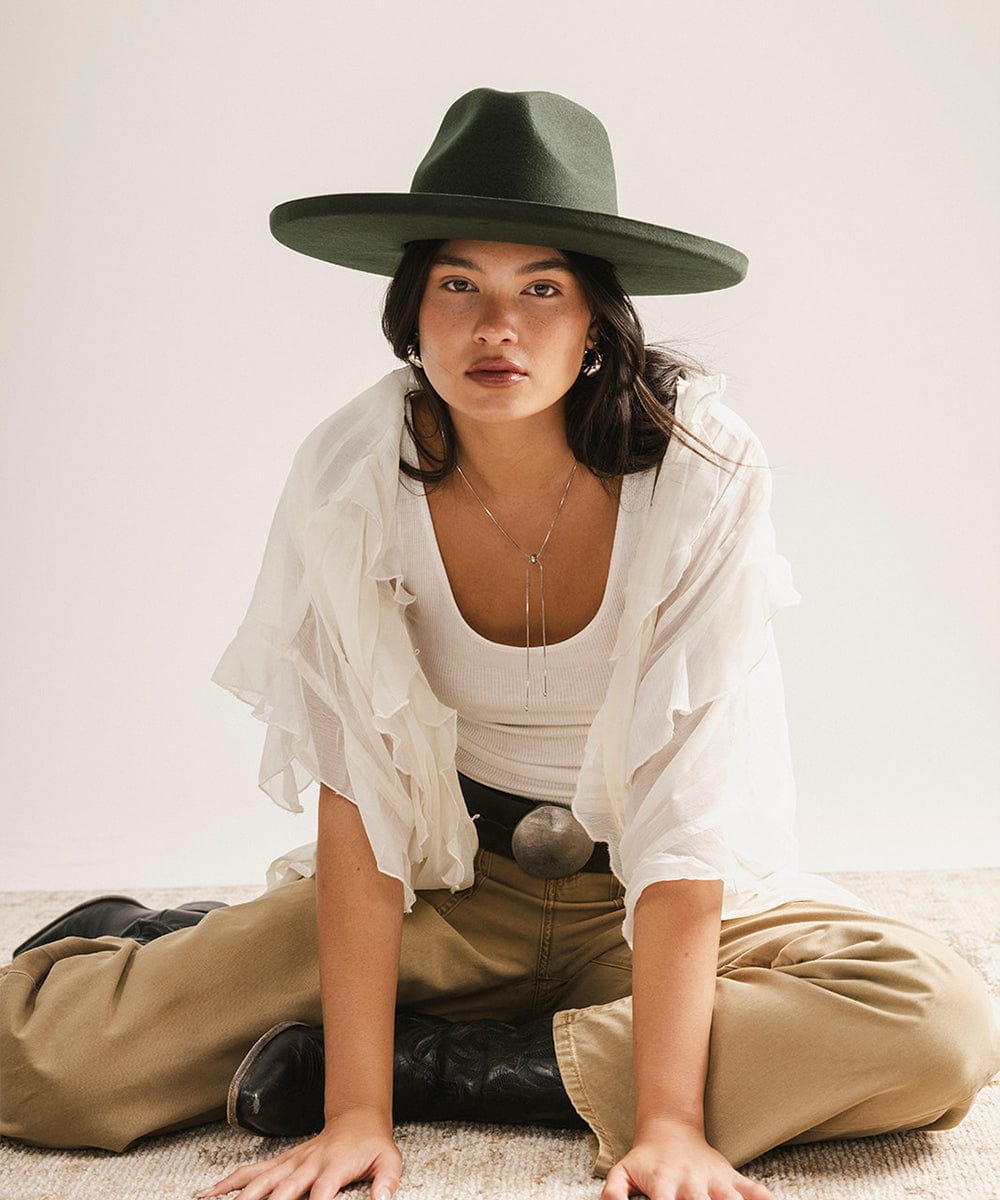 Woman wearing a dark green hat, white top, beige pants, and black boots sitting on the floor against a white background. #color_dark green