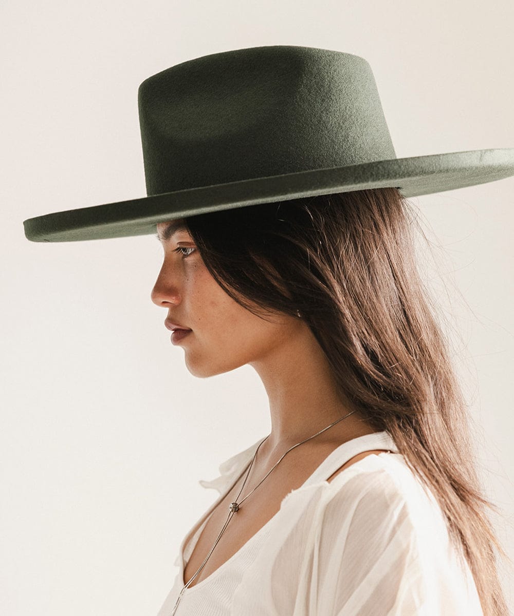 Woman wearing a dark green pencil brim fedora hat and a white top, facing sideways against a plain background #color_dark green