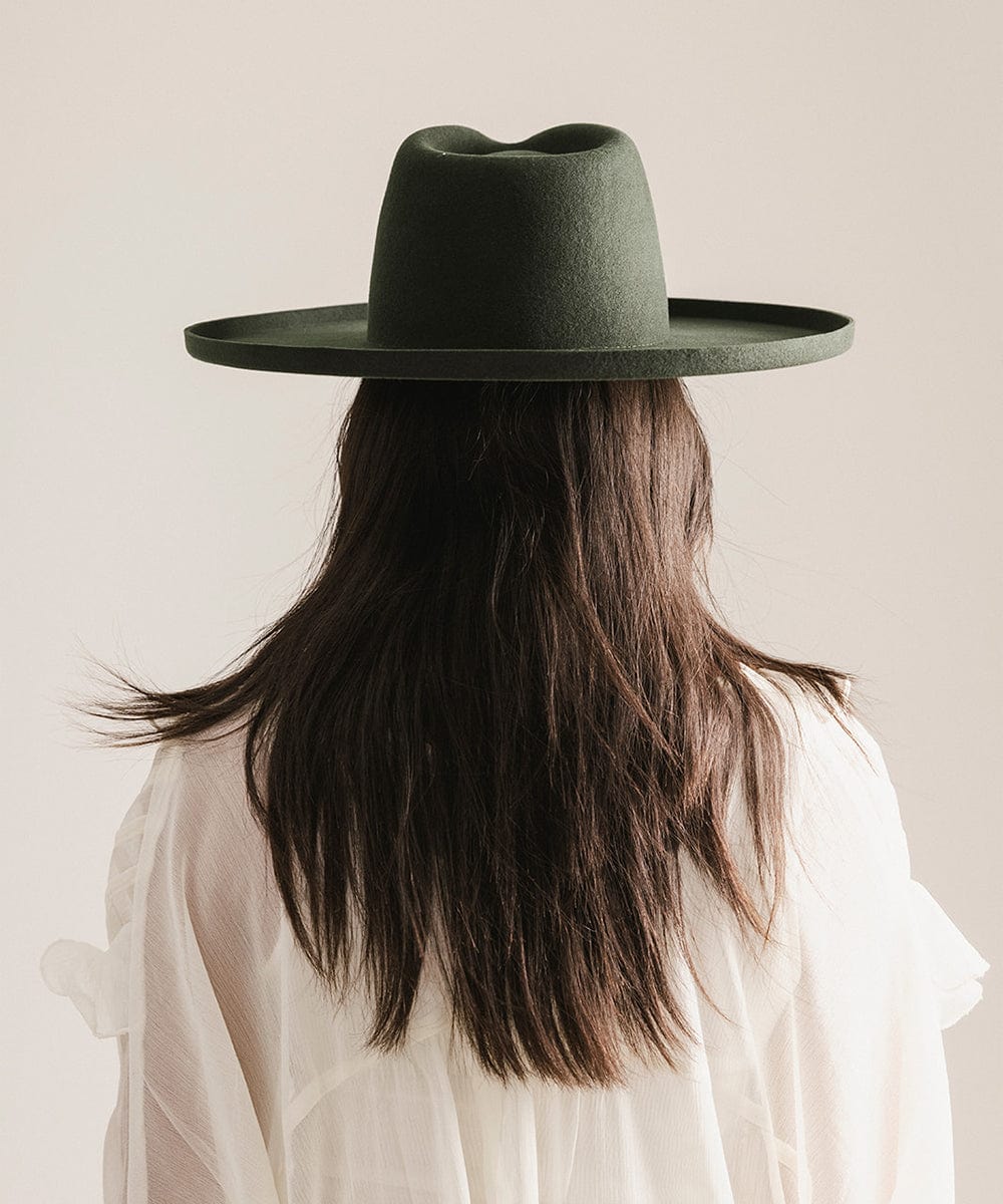 Woman wearing a dark green pencil brim fedora hat and a white top, facing behind against a plain background #color_dark green