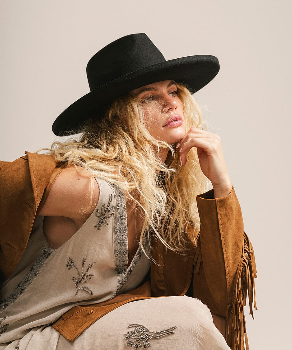 Woman wearing a black hat and a light colored dress with a brown jacket, facing sideways against a plain background #color_black